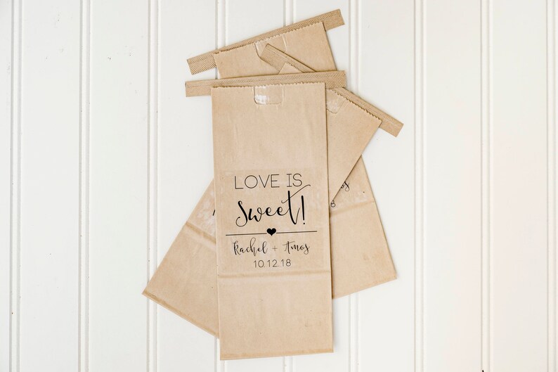 May include: Three brown paper bags with a white label on the front. The label says "Love is Sweet!" with a small heart and the text "Rachel + Thomas 10.12.18".