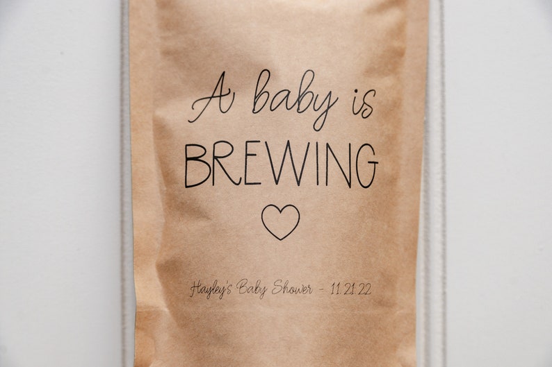Pu&ograve; includere: Un sacchetto di carta marrone con il testo "A baby is BREWING" e un cuore. Il sacchetto ha anche il testo "Hayley's Baby Shower - 11.21.22".