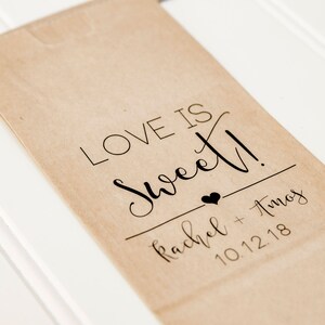 May include: A brown paper bag with the text "Love is Sweet!" printed on it. Below the text is a small heart and the names "Rachel + Amos" and the date "10.12.18".
