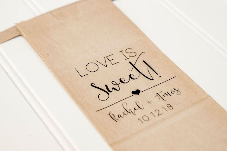 May include: A brown paper bag with the text "Love is Sweet!" printed on it. Below the text is a small heart and the names "Rachel + Amos" and the date "10.12.18".