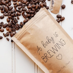 Pu&ograve; includere: Un sacchetto di carta marrone con il testo "A baby is BREWING" e un cuore. Il testo "Ashley's Baby Shower - 11/21/22" &egrave; anche sul sacchetto. I chicchi di caff&egrave; sono sparsi intorno al sacchetto.