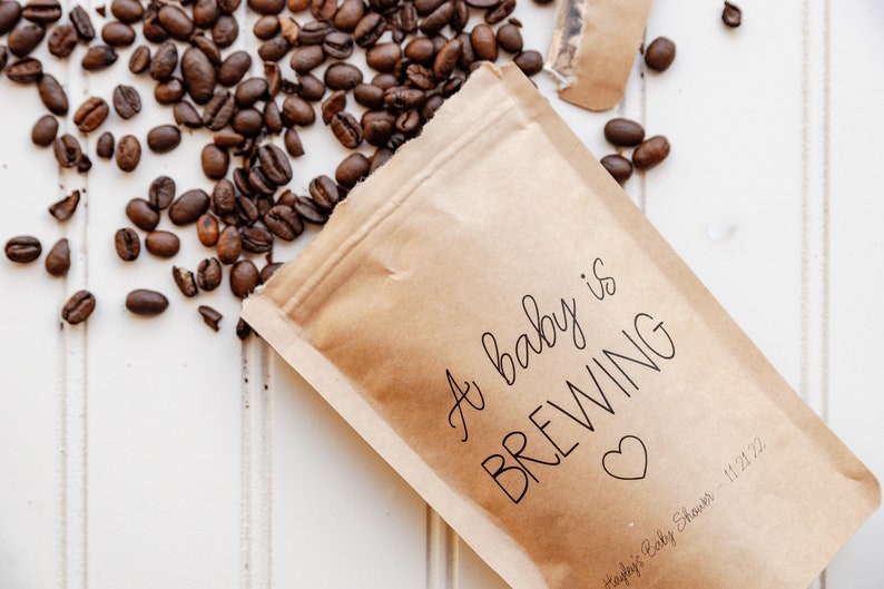 Pu&ograve; includere: Un sacchetto di carta marrone con il testo "A baby is BREWING" e un cuore. Il testo "Ashley's Baby Shower - 11/21/22" &egrave; anche sul sacchetto. I chicchi di caff&egrave; sono sparsi intorno al sacchetto.