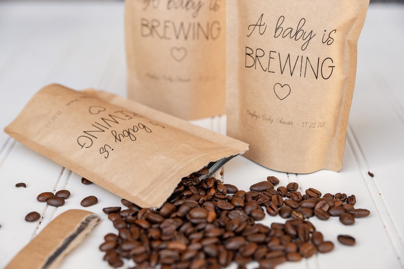 Pu&ograve; includere: Tre sacchetti di carta marrone con la scritta "A baby is brewing" e un cuore stampato su di essi. I sacchetti sono pieni di chicchi di caff&egrave;, che stanno fuoriuscendo da uno dei sacchetti.