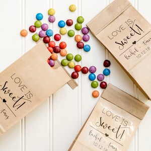 May include: Three brown paper bags with the text "LOVE IS Sweet!" and the names "Rachel + Tomos" and the date "10.12.18". The bags are filled with colorful candy.