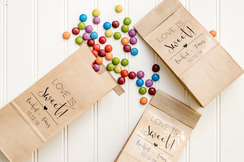 May include: Three brown paper bags with the text "LOVE IS Sweet!" and the names "Rachel + Tomos" and the date "10.12.18". The bags are filled with colorful candy.