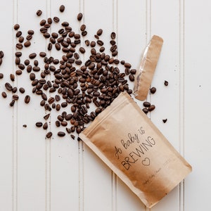 Pu&ograve; includere: Un sacchetto di carta marrone con il testo "A baby is brewing" e un piccolo cuore. I chicchi di caff&egrave; si riversano dalla borsa su una superficie bianca.