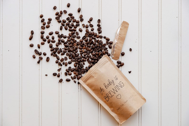 Pu&ograve; includere: Un sacchetto di carta marrone con il testo "A baby is brewing" e un piccolo cuore. I chicchi di caff&egrave; si riversano dalla borsa su una superficie bianca.