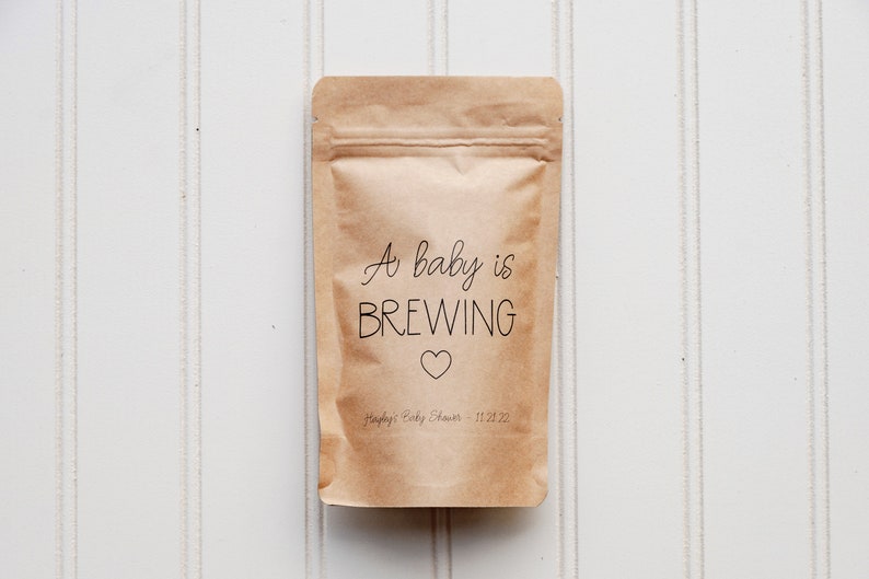 Pu&ograve; includere: Un sacchetto di carta marrone con un'etichetta bianca su cui &egrave; scritto "A baby is BREWING" con un piccolo cuore sotto il testo. L'etichetta riporta anche "Happy's Baby Shower 11.21.22".