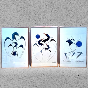 May include: Three framed art prints with abstract designs of birds and a deer in blue and black. Each print is in a gold frame, set against a light blue backdrop. The artwork has a minimalist, abstract style, with each piece signed.