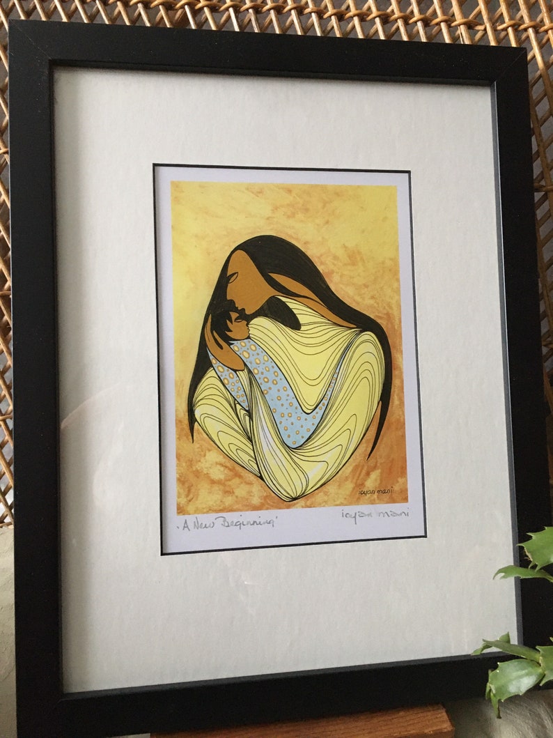 May include: A framed print of a Native American art piece depicting a mother and child. The mother is holding the child close, and both are surrounded by yellow and blue swirls. The title of the piece is "A New Beginning" and is signed by the artist, "Joya Mani".