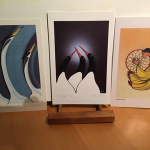 May include: Three art prints displayed on a wooden easel. The prints feature stylized bird and Native American-themed illustrations. One print shows three birds with yellow beaks, another depicts two birds with red beaks, and the third shows a figure with a dreamcatcher.