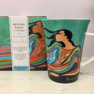 May include: A turquoise ceramic mug with a colourful abstract design featuring a woman's face and swirling lines. The mug has a white handle and the text "Maxine Noel" is printed on the bottom. The mug is accompanied by a box with the text "Mother Earth" and "La Terre-Mere" printed on it.