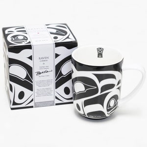 Könnte beinhalten: Schwarze und weiße Keramiktasse mit einem Rabenmotiv. Die Tasse ist Teil eines Sets von Tassen mit verschiedenen indigenen Kunstwerken. Die Schachtel zeigt die Unterschrift des Künstlers und die Worte "Raven Corbeau by."