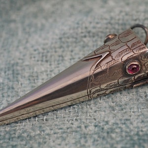 May include: A silver-toned, snake-shaped pendant with a pointed end. The head features engraved scales and a red gemstone eye. A small ring is attached to the head for hanging. The pendant is likely made of metal.