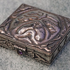 May include: A silver-colored, rectangular box with intricate embossed designs on the lid and sides. The lid features a central, raised design. A small, purple gemstone is set into the clasp. The box is likely used for jewelry or small keepsakes.