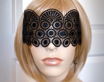 May include: Black lace eye mask with a scalloped edge and circular cutouts. The mask is worn on a mannequin with blonde hair.