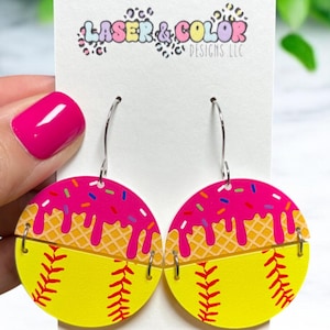 May include: A pair of softball-themed earrings. The earrings are yellow with red stitching, and topped with pink frosting and sprinkles. The earrings are attached to silver hooks. The text "LASER & COLOR DESIGNS LLC" is on the backing card.