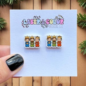 May include: A pair of earrings featuring three kings wearing crowns, in blue, gold, and red. The earrings are on a white card with the text "LASER & COLOR DESIGNS LLC". The background includes Christmas decorations and a wooden surface.