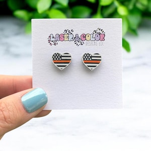May include: A pair of heart-shaped earrings with a thin red line across the center of a white and black American flag design. The earrings are displayed on a white card with the text "Laser & Color Designs LLC".