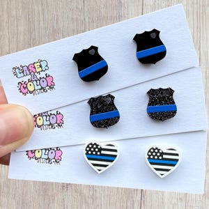 May include: Three pairs of earrings on a white card. The earrings feature a black police badge design with a blue stripe, a glittery black badge with a blue stripe, and a white heart with a blue stripe design. The card has the text "LASER COLOR DESIGNS".
