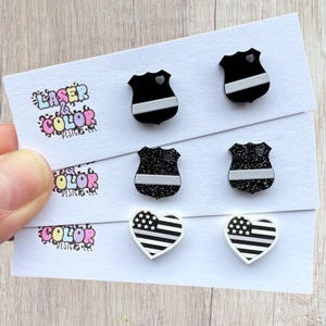 May include: A selection of stud earrings on white cards. The earrings feature shield and heart shapes with black and white designs, including a thin blue line and an American flag pattern. The cards have the text "Laser Color Designs".