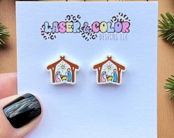 May include: A pair of Christmas-themed stud earrings featuring a nativity scene inside a wooden stable. The earrings are on a white card with the text "LASER & COLOR DESIGNS LLC". The background includes pine branches, pine cones, and red berries.