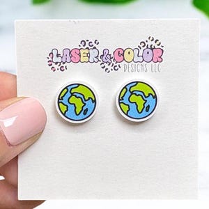 May include: A pair of round stud earrings featuring a blue outline and a green and yellow Earth design. The earrings are on a white card with the text "LASER & COLOR DESIGNS LLC". The earrings are held by a person.