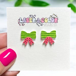 May include: A pair of watermelon bow earrings on a white card. The earrings have green striped bows with red and black watermelon details. The card has the text "LASER & COLOR DESIGNS LLC" in colorful letters.