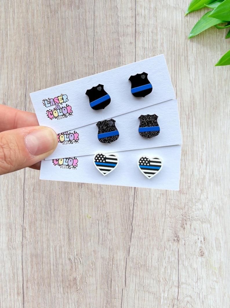 May include: Three pairs of earrings on a white card. The earrings feature a black police badge design with a blue stripe, a glittery black badge with a blue stripe, and a white heart with a blue stripe design. The card has the text "LASER COLOR DESIGNS".