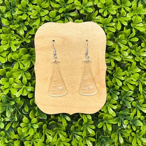 May include: A pair of clear acrylic earrings shaped like beakers with silver hooks. The earrings are displayed on a wooden stand against a green background.