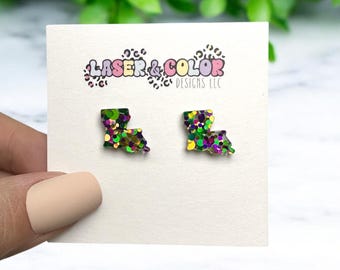 May include: A pair of earrings shaped like the state of Louisiana, covered in green, purple, and gold glitter. The earrings are on a white card with the text "Laser & Color Designs LLC".