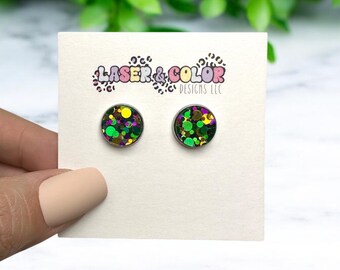 May include: A pair of round stud earrings with a black background and green, gold, and purple glitter. The earrings are displayed on a white card with the text "Laser & Color Designs LLC".
