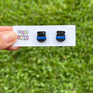May include: Two black glitter shield-shaped earrings with a blue stripe. The earrings are presented on a white card with the text "Laser & Color Designs". The background is blurred green grass.