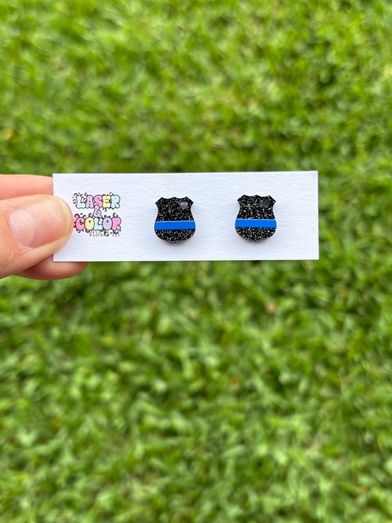 May include: Two black glitter shield-shaped earrings with a blue stripe. The earrings are presented on a white card with the text "Laser & Color Designs". The background is blurred green grass.