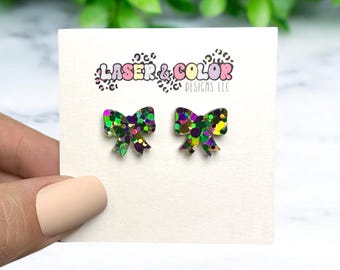 May include: A pair of small, glittery bow earrings in green, purple, gold, and pink. The earrings are on a white card with the text "Laser & Color Designs LLC".