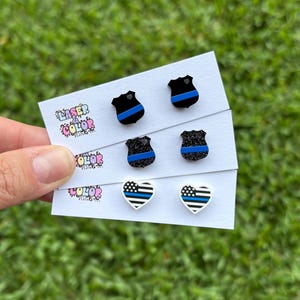 May include: A selection of stud earrings displayed on white cards. The earrings showcase shield and heart shapes with blue and black patterns. The cards display the text "Laser Color Designs" in colourful lettering. The background is green grass.