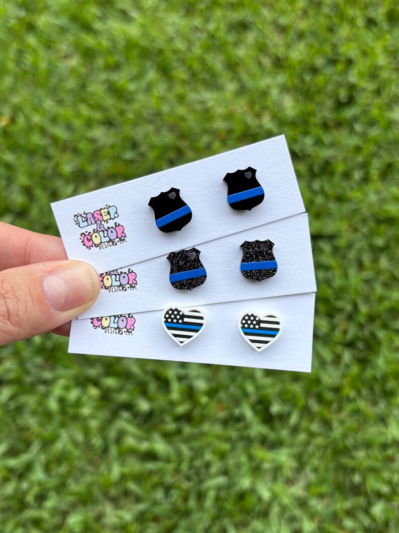 May include: A selection of stud earrings displayed on white cards. The earrings showcase shield and heart shapes with blue and black patterns. The cards display the text "Laser Color Designs" in colourful lettering. The background is green grass.