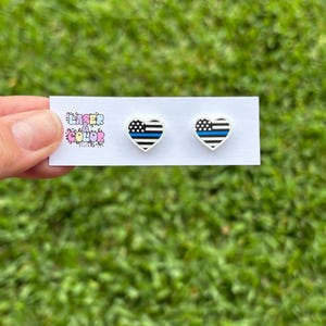 May include: Heart-shaped stud earrings with a black and white striped design and a blue line, resembling a flag. The earrings are on a white card with the text "Laser Color Designs". The background is green grass.