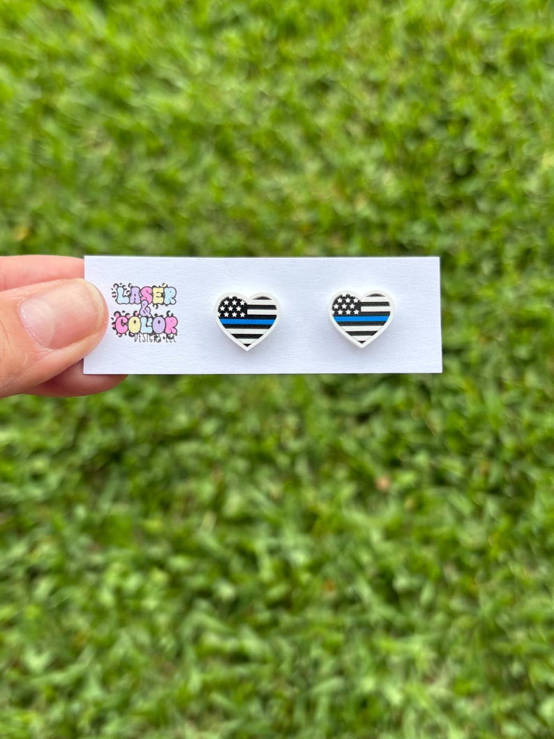 May include: Heart-shaped stud earrings with a black and white striped design and a blue line, resembling a flag. The earrings are on a white card with the text "Laser Color Designs". The background is green grass.