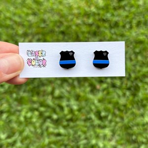 May include: A pair of black shield-shaped stud earrings with a blue stripe and a small heart detail. The earrings are displayed on a white card with the text "Laser & Color Designs". The background is blurred green grass.