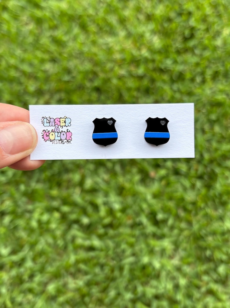 May include: A pair of black shield-shaped stud earrings with a blue stripe and a small heart detail. The earrings are displayed on a white card with the text "Laser & Color Designs". The background is blurred green grass.