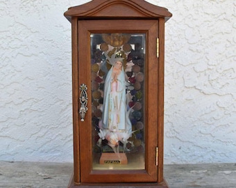 Può includere: Una teca in legno con una parte anteriore in vetro, che presenta una statua della Vergine Maria. La statua è vestita di bianco e blu, e la teca è decorata con perline colorate e la parola "FATIMA".