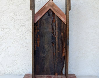 May include: A rustic wooden sculpture with a triangular roof made of weathered wood and copper. The sculpture is standing on a wooden base.