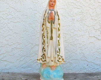 Puede incluir: Estatua blanca de la Virgen María, Nuestra Señora de Fátima, con una túnica blanca y ribetes dorados, de pie sobre una base de nube azul. La estatua sostiene un rosario en sus manos y tiene una corona en la cabeza.