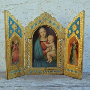 Może przedstawiać: Złoty drewniany tryptyk z centralnym panelem przedstawiającym Matkę Boską trzymającą Dzieciątko Jezus. Panele boczne przedstawiają anioły z skrzydłami. Tryptyk jest ozdobiony niebieskimi i złotymi akcentami.