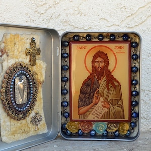May include: A small, silver tin box with a blue and gold rosary inside. The box is open and the rosary is visible. The rosary is made of blue beads and gold accents. The box also contains a small, framed image of Saint John the Forerunner. The image is surrounded by a gold frame and has a gold background. The image shows Saint John the Forerunner with a long, brown beard and hair. He is wearing a brown robe and holding a scroll in his hand. The scroll has text on it that reads "Repent for the Kingdom of Heaven is at Hand."