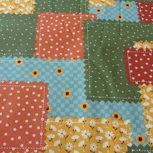 May include: A patchwork fabric with a variety of floral and geometric patterns in shades of yellow, green, blue, and orange. The fabric is made up of squares with scalloped edges.