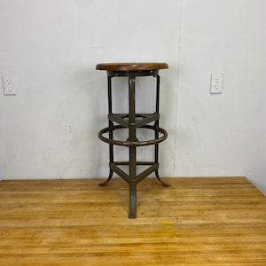 May include: A vintage industrial stool with a wooden seat and a metal frame. The stool has a footrest and a triangular base.