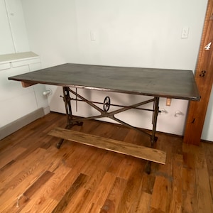 May include: A vintage industrial-style drafting table with a wooden top and metal base. The table has a crank mechanism for adjusting the height and a wooden footrest.