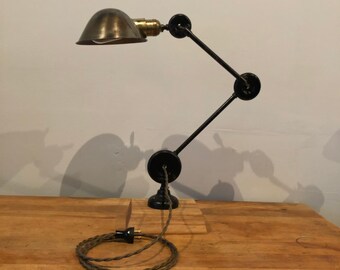 May include: A black adjustable desk lamp with a brass shade and a green braided cord. The lamp is on a wooden base and has a plug.